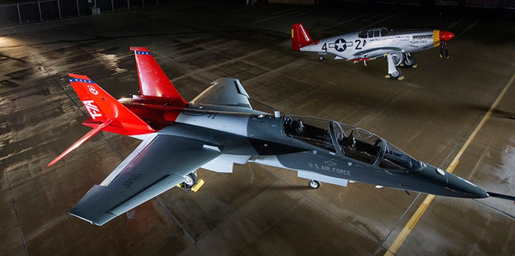 A gray and red Boeing T-7A Red Hawk jet trainer parked in a hangar, with a restored P-51C Mustang with a red tail behind it